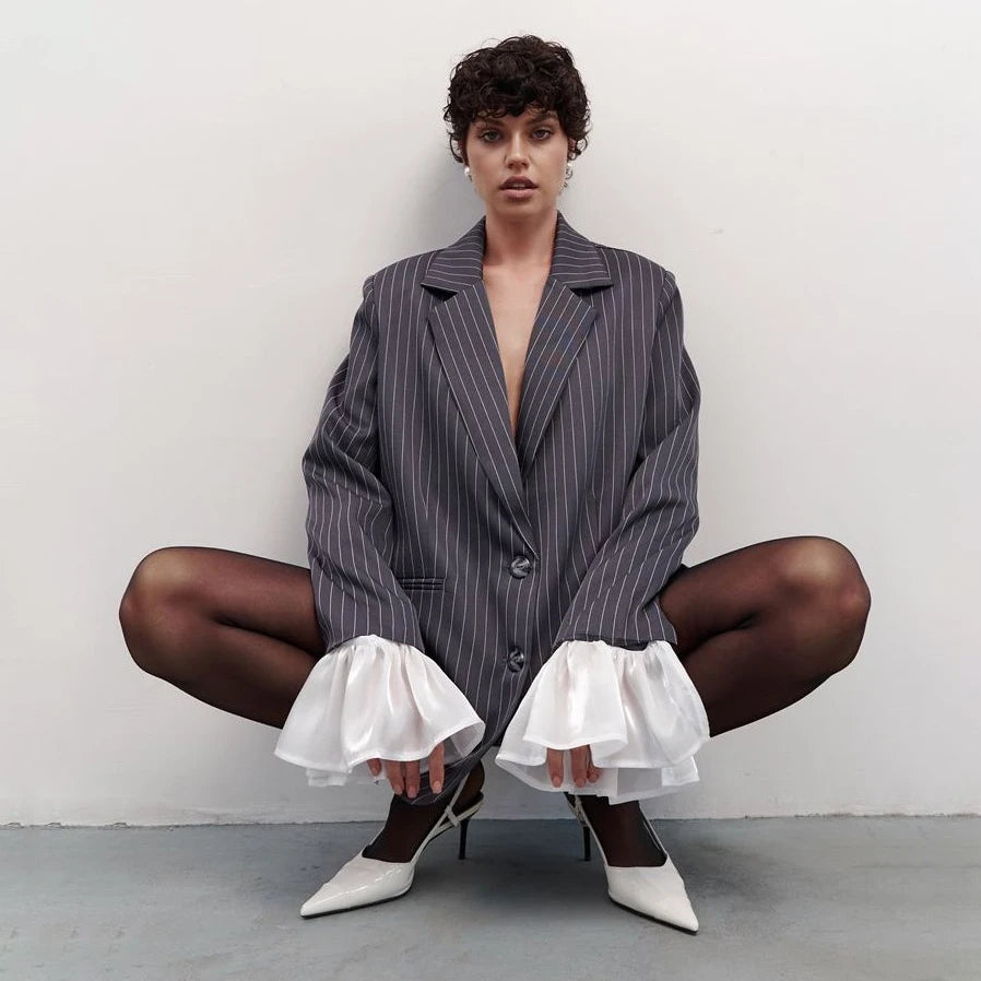 Person wearing a pinstripe blazer and white pants with ruffles, sitting on the floor against a plain background.